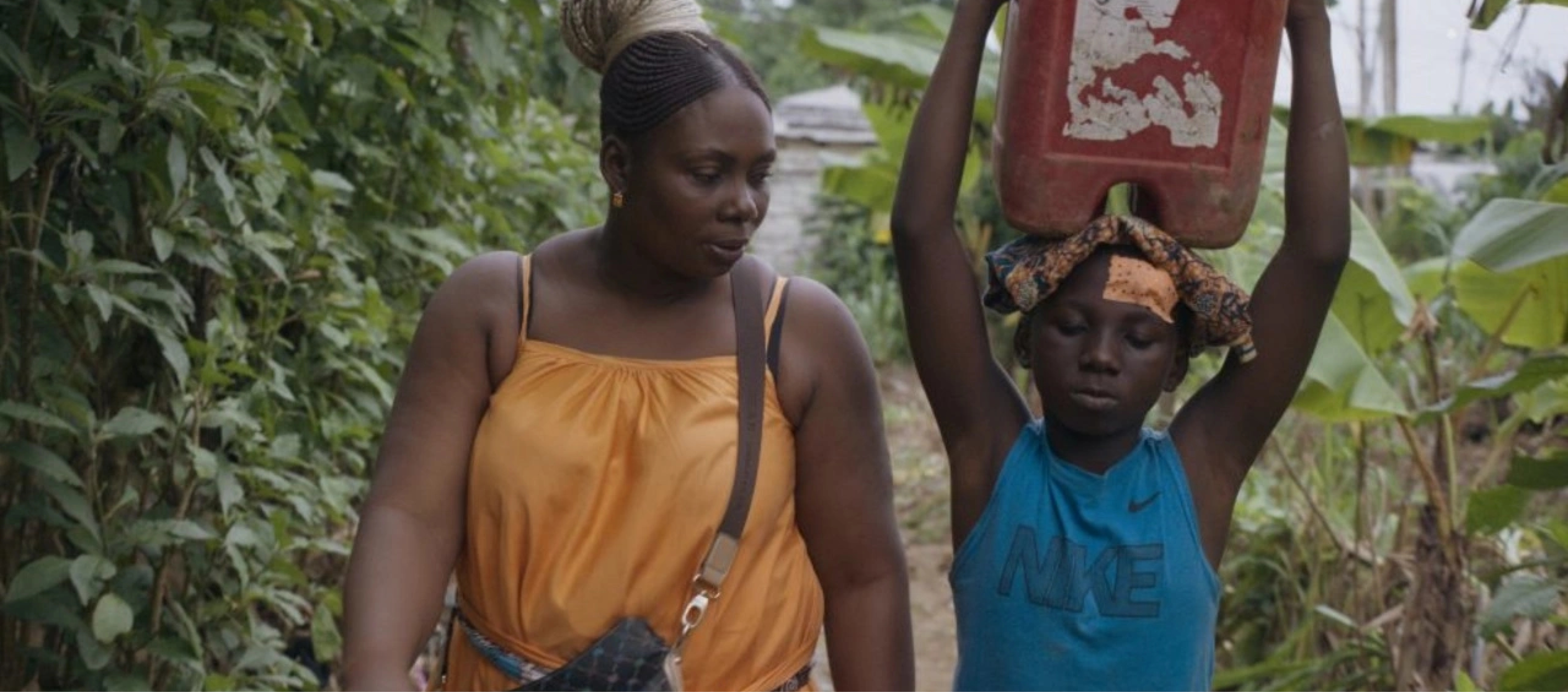 A Cameroonian woman wearing an orange dress walks on a path through a lush landscape next to a child who is holding a large red canister atop their head.