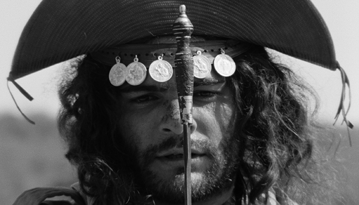 A bearded bandit with long curly hair, holds the hilt of a sword in front of his face, while wearing a large-brimmed hat adorned with coins. 