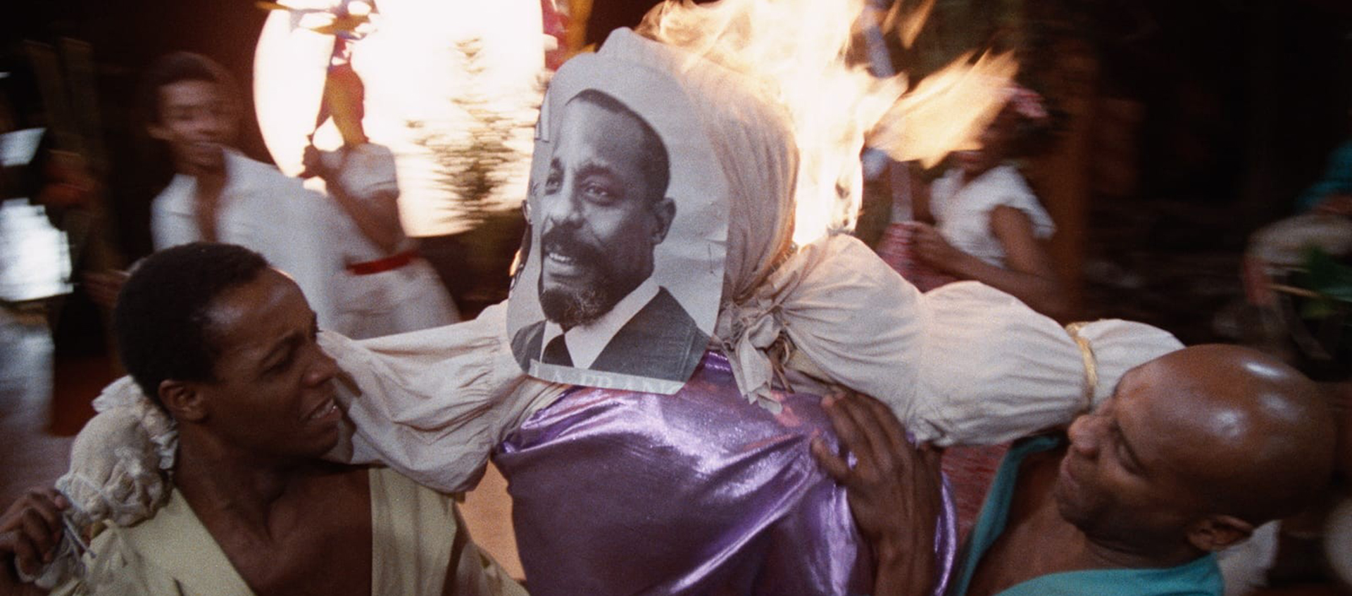 Two Black people hold up an effigy that is on fire, a photo of a Black man is attached to the face of the effigy.