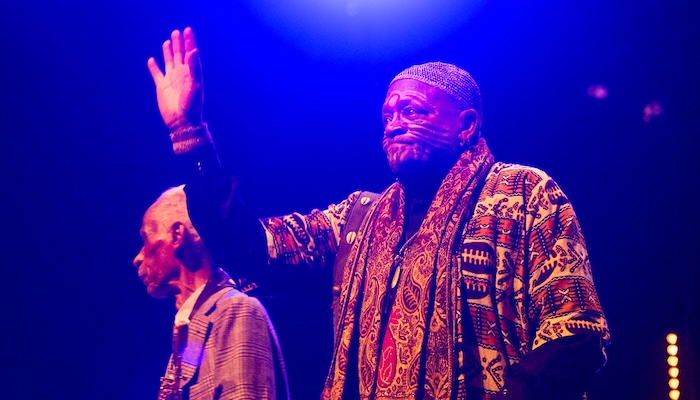 Two older men stand on stage under a blue light, one is wearing tribal face paint and waiving.  