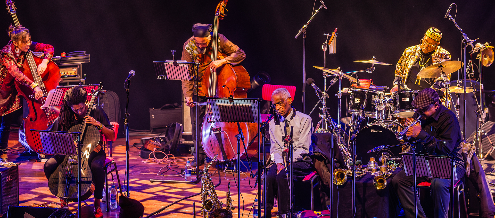 Six jazz musicians perform onstage, playing upright bass, drums, and the trumpet. Roscoe Mitchell is seated center stage, and Don Moye plays drums. 