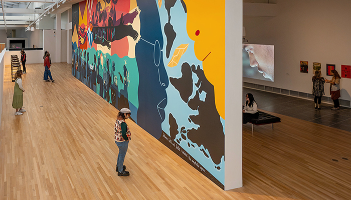 A large colorful mural painted on a long wall in a brightly lit gallery, with a video projection and posters in a darkened space in the background.